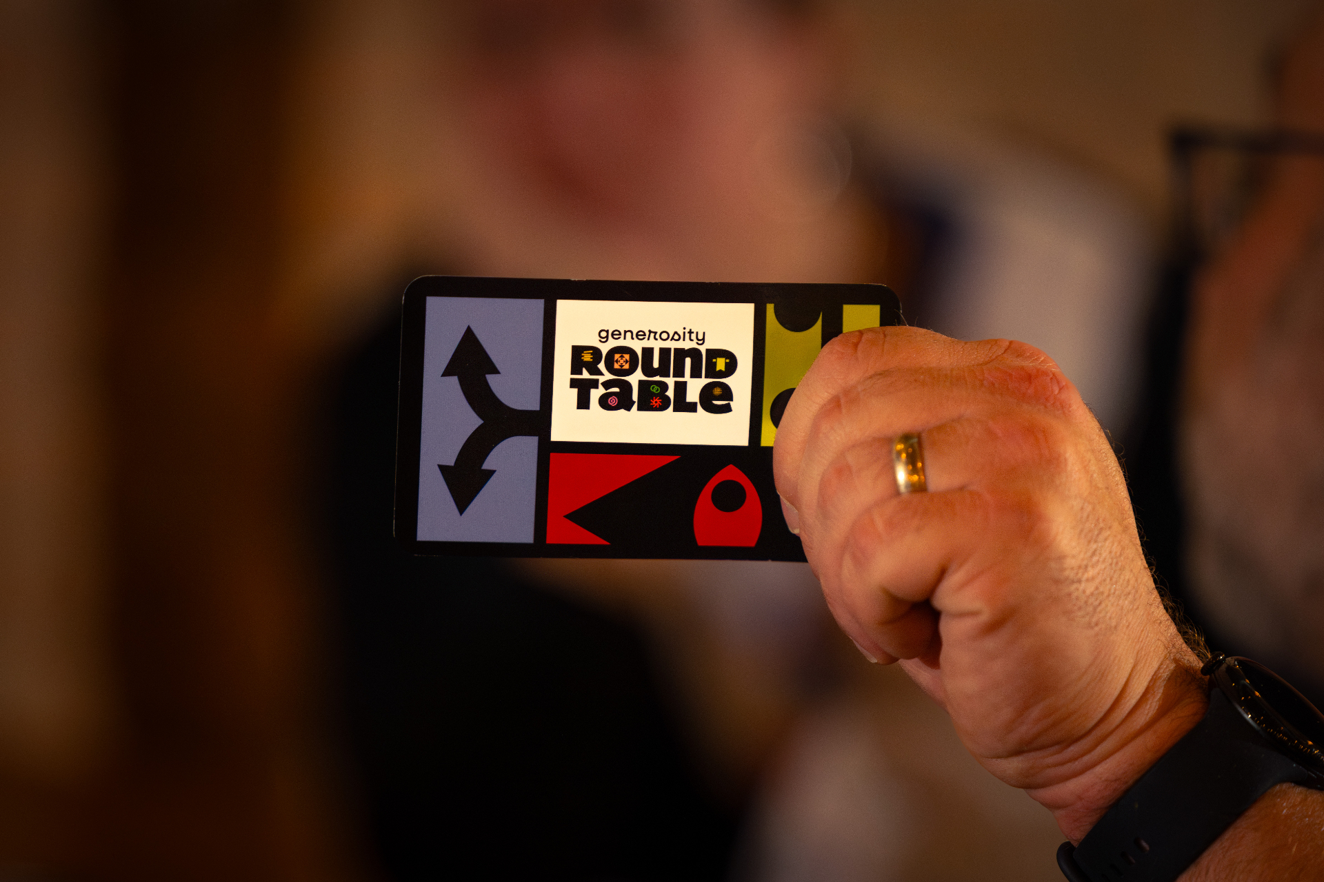 A hand holding a Generosity Roundtable sticker, with cards blurred in the background.